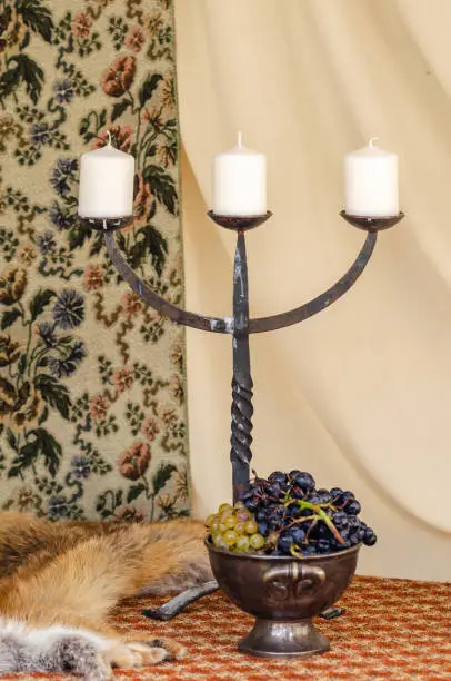 Medieval three-armed candlestick, bowl with grapes and fur coat from a dead fox. Still life. Vertical photo