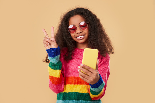 Happy African American hipster girl in colorful knitted sweater and stylish sunglasses showing peace sign and taking selfie with smartphone while standing against beige background