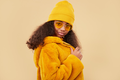 Side view of African American kid in yellow hat and sunglasses adjusting stylish fur coat and looking at camera on beige background
