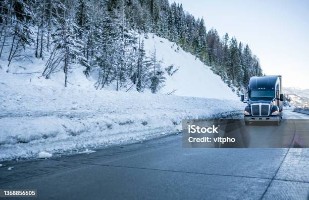 Black Big Rig Bonnet Industrial Semi Truck Transporting Cargo In Semi Trailer Running On The Winding Winter Dangerous Slippery Road With Snow And Ice And With Snowy Trees On The Hill Side Stock Photo - Download Image Now
