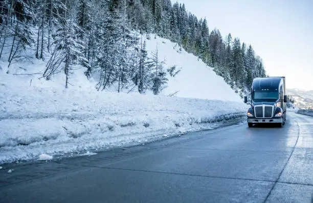 Photo of Black big rig bonnet industrial semi truck transporting cargo in semi trailer running on the winding winter dangerous slippery road with snow and ice and with snowy trees on the hill side