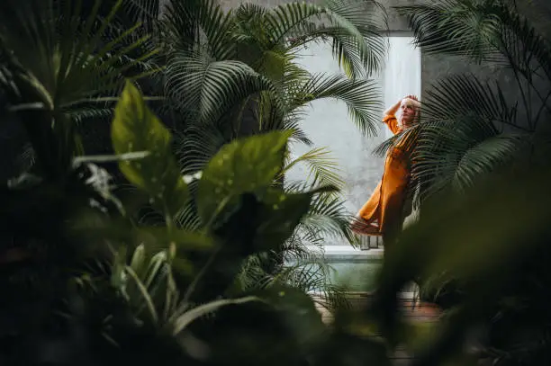 Photo of Woman leaning on a gray concrete wall surrounded by green tropical palm leaves, by the pool.