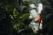 Woman leaning on a gray concrete stone wall surrounded by green tropical palm leaves, by the pool.