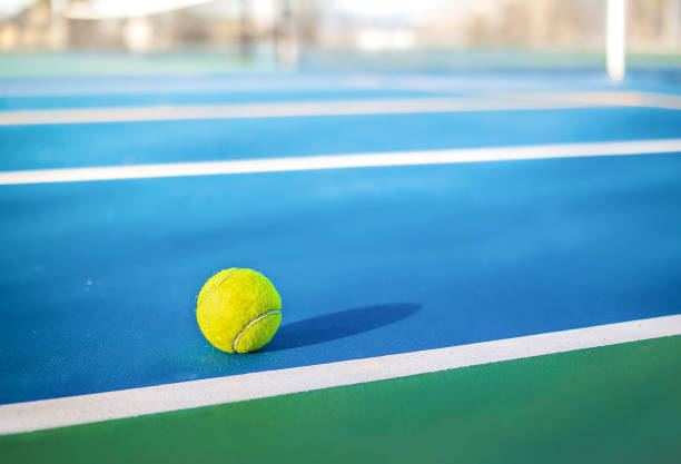 Yellow tennis ball next to sideline in outdoor tennis court, closeup. Defocused and abstract blue and green rubberized ground surface for shock absorption. Tennis sport texture. Selective focus. rubberized stock pictures, royalty-free photos & images