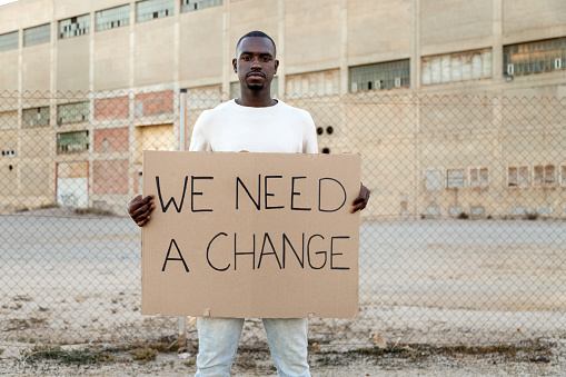 Young African American man looking at camera holding a we need a change banner protesting for protests movement. Copy space. Social movement activist concept.