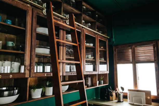 Classic kitchen interior with high brown coloured wooden cabinets and ladder fully equipped with kitchen utensils against dark green coloured wall. Sunlight coming through window