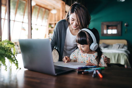 Young Asian mother homeschooling her daughter and assist her with school work at home. Little girl with headphones is studying from home and attending online school classes with laptop. E-learning, homeschooling concept
