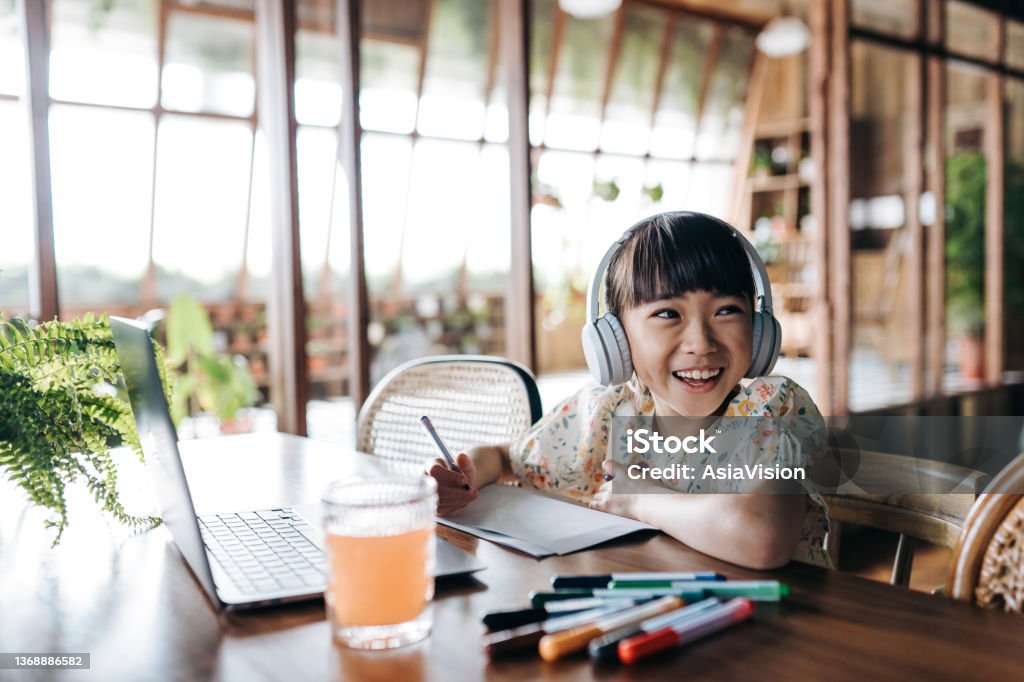 Cheerful Asian girl with headphones studying from home, smiling joyfully. She is attending online school classes with laptop and writing notes at home. E-learning, homeschooling concept Child Stock Photo