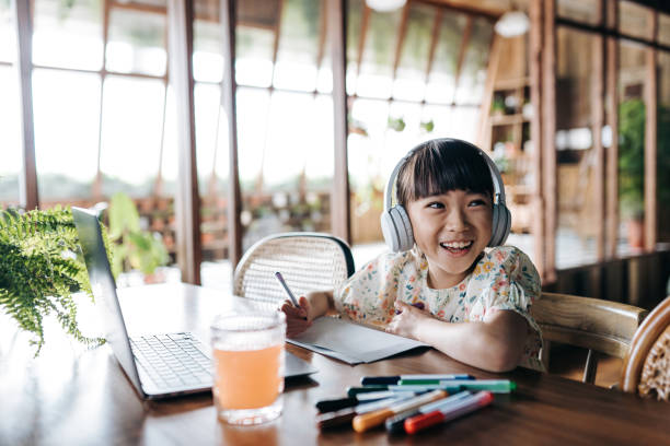 cheerful asian girl with headphones studying from home, smiling joyfully. she is attending online school classes with laptop and writing notes at home. e-learning, homeschooling concept - chinese ethnicity audio imagens e fotografias de stock