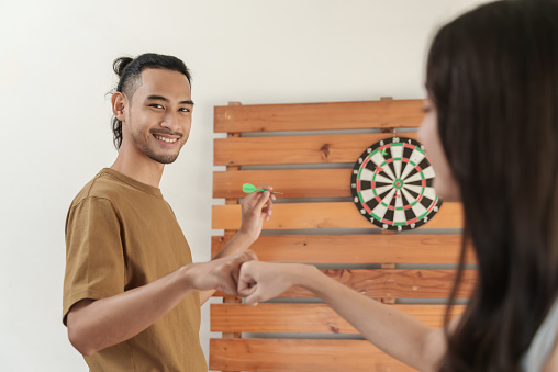 Young Asian couple enjoy to playing darts together. Young man smiling and looking at camera while playing darts and making fist bump with his friend.