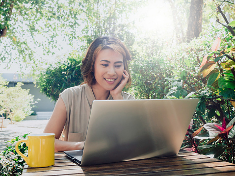 Attractive Asian female portrait with short hair sitting with a hand under chin, enjoy with coffee and looking at the laptop computer screen with a relaxing smile in the outdoor garden.