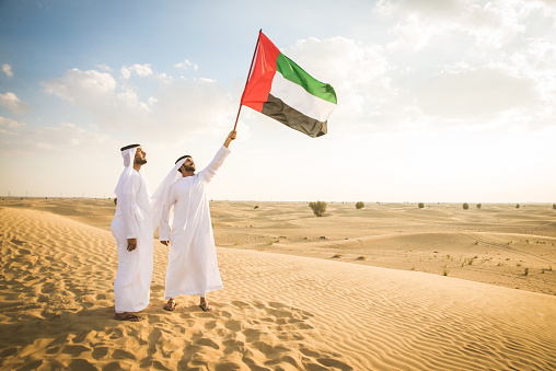 Arabian men with kandora walking in the desert and holding emirates national flag- Portrait of two middle eastern adults with traditional arabian dish dash dress