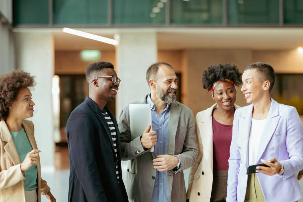 Winning Team Group of Diverse Coworkers Standing at the Office Corridor and Smiling while Having an Informal Conversation professional people laughing stock pictures, royalty-free photos & images
