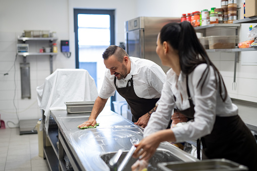 A chef and cook cleaning the workspace after doing dishes indoors in restaurant kitchen.