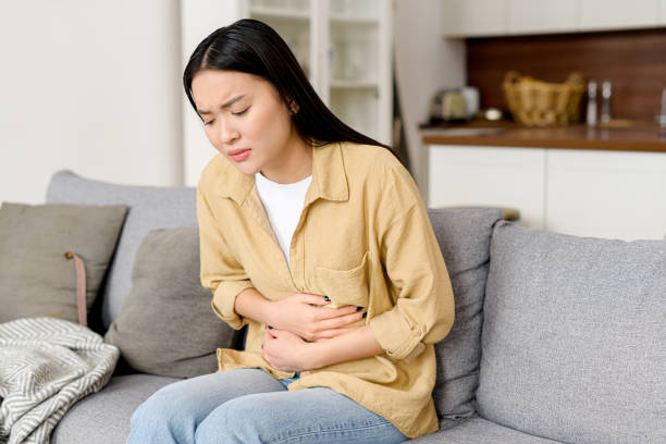 Concerned asian woman suffering from stomach-ache sitting on sofa. Female holding tummy, undergoing belly pain and discomfort stock photo