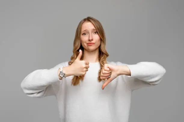 Puzzled blonde girl showing thumb up and down. Skeptical thinking woman judging deciding how to rate a situation, wears white sweater, gray background