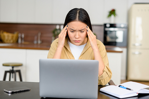 Tired and worn out from online work young woman sitting in front laptop holding head, feels strong headache. Female freelance with eyes closed is massaging head temples with suffering face expression