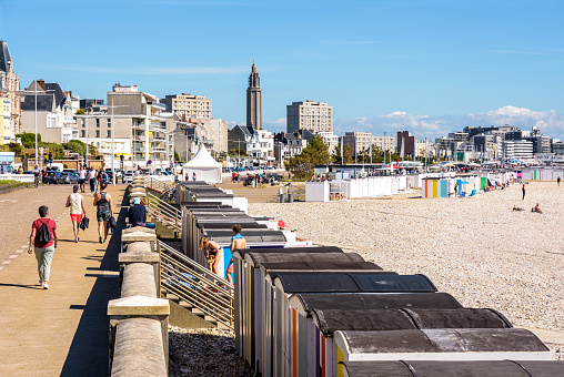 Le Havre, France - June 11, 2021: People are strolling along the pebble beach lined with private huts, with the bell tower of St. Joseph's church in the distance, on a sunny day.
