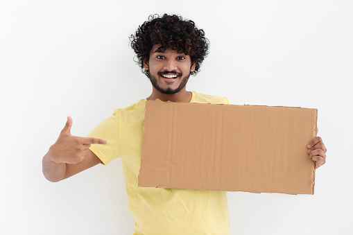 Happy Indian man with curly hair in yellow t-shirt holding and pointing at an empty cardboard sign looking at the camera and smiling on white background. Job search, sale concept