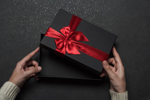 istock Hands in a sweater opening a black gift box with a red bow on a black granite background. 1368937502