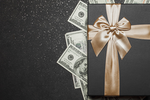 Black gift box with gold ribbon and a big bow on a pile of money. Gift on a granite surface.