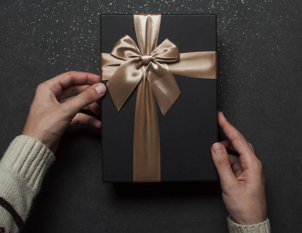 istock Hands in a sweater opening a black gift box with a golden bow on a black granite background. 1368937505