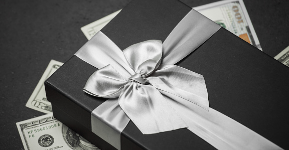 A black gift box with a silver ribbon and a large bow on a pile of money. Gift on a granite surface.