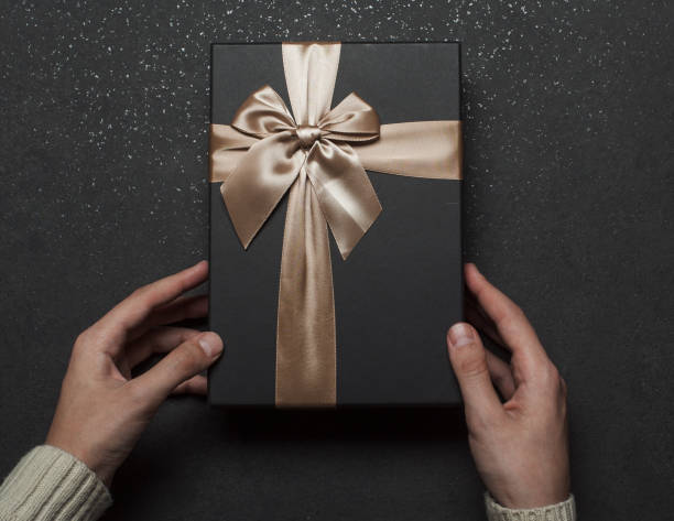 istock Hands in a sweater holding a black gift box with a golden bow on a black granite background. 1368937525
