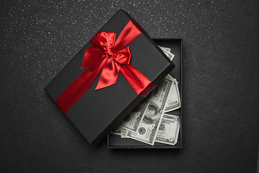 An open black gift box with a red ribbon and a large bow with money inside. Granite background