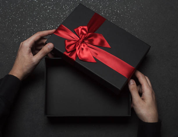 istock The hands of a guy in a black shirt open a gift box with a large red bow. Empty box. Granite background. 1368937756