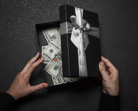 The hands of a guy in a black shirt open a gift box with a large silver bow. Money box. Granite background.