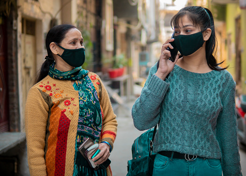 Indian matures mother and young daughter walking together on the street and taking on a smartphone. They are wearing protective face mask to prevent coronavirus. COVID -19 pandemic.