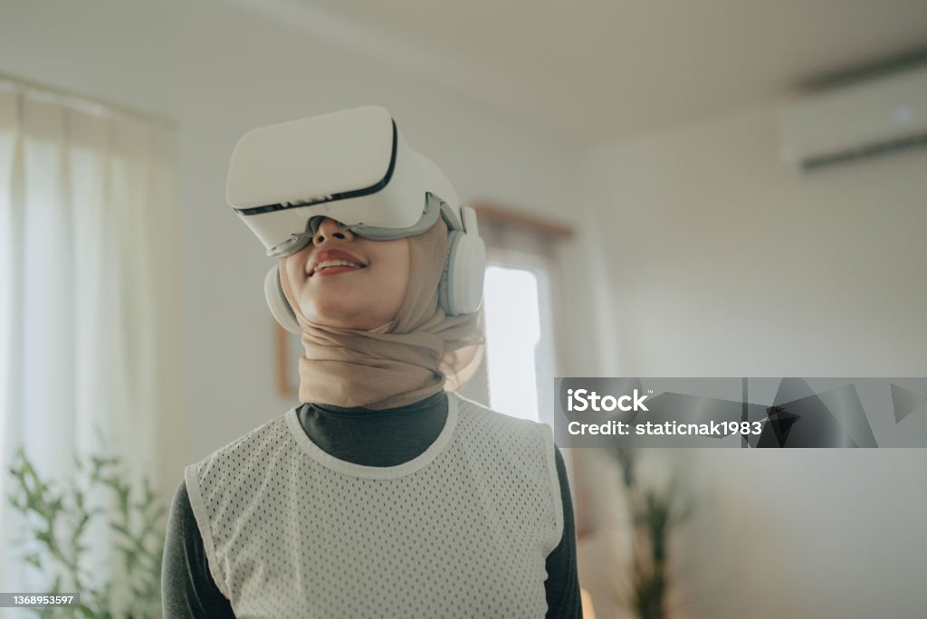 Attractive islam woman wearing VR Glasses sitting on sofa at home. Young woman uses a virtual reality glasses control panel. Girl with pleasure uses head-mounted display. Asian woman playing game in virtual reality glasses. Virtual Reality Simulator Stock Photo