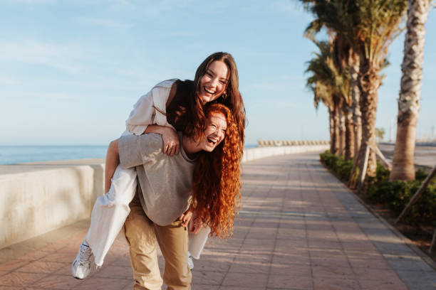 beztroska młoda kobieta piggybacking swojego przyjaciela na promenadzie - young adult beach people cheerful zdjęcia i obrazy z banku zdjęć