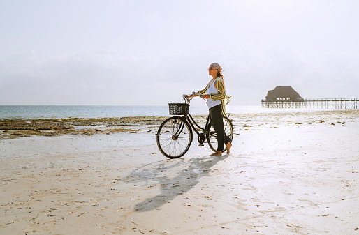 Female dressed light summer clothes have morning walk with old vintage bicycle with front basket on the lonely low tide ocean white sand coast on Kiwengwa beach on Zanzibar island, Tanzania.