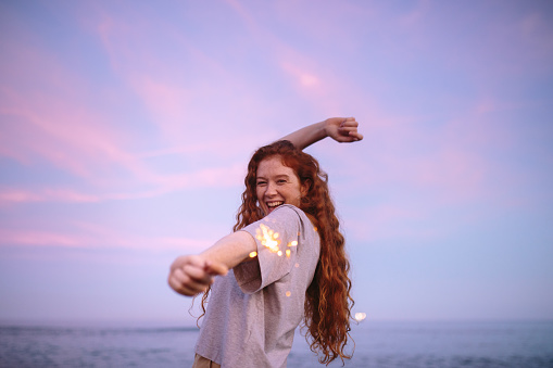 Lighting things up at the beach. Happy young woman dancing and laughing while holding a sparkling light in her hand. Cheerful young woman celebrating with bengal lights at the beach.