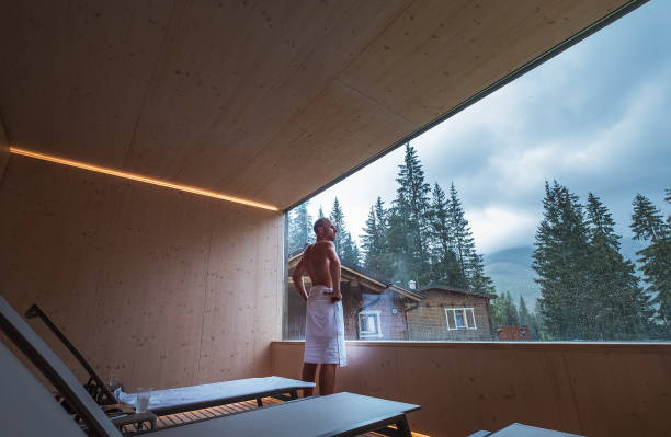 Relaxing man wrapped white sheet after Finnish sauna in Tepidarium with wide window enjoying the green forest view and pleasant healthy body care temperature treatment. Body healthcare concept image. relajante hombre envuelto en sábanas blancas después de la sauna finlandesa en tepidarium con amplia ventana disfrutando de la vista verde del bosque y agradable tratamiento de temperatura de cuidado corporal saludable. imagen conceptual de cuidado de la