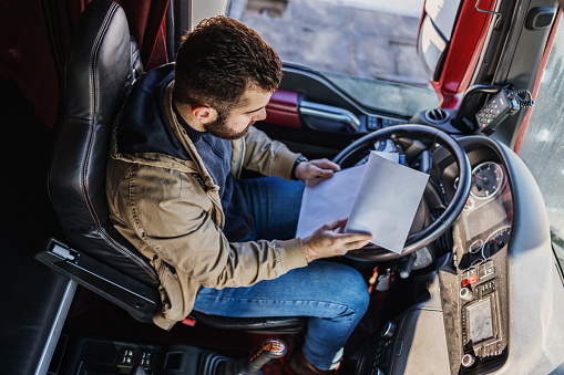 Young bearded man reading some papers and tour documents before he starts with a new driving tour. View from above inside of truck cabin.