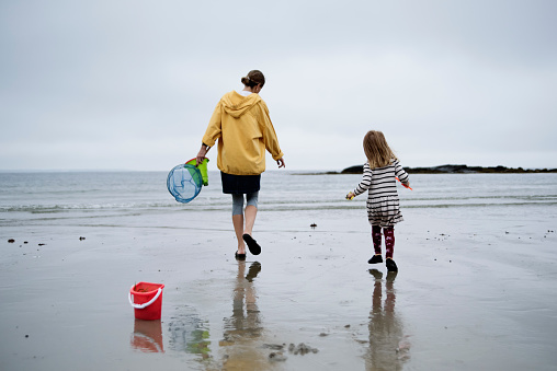 Mother and 4 year’s old daughter looking for seashells on beach on a chilly and cloudy summer day. Mom is wearing a yellow hoodie and little girl a striped dress. Horizontal full length outdoors shot with copy space.
