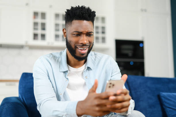 istock Focused bearded multiracial man sitting on the sofa at home and holding smartphone. Guy reading news online, spending leisure time in social networks, dark-haired male texting and chatting 1368977395