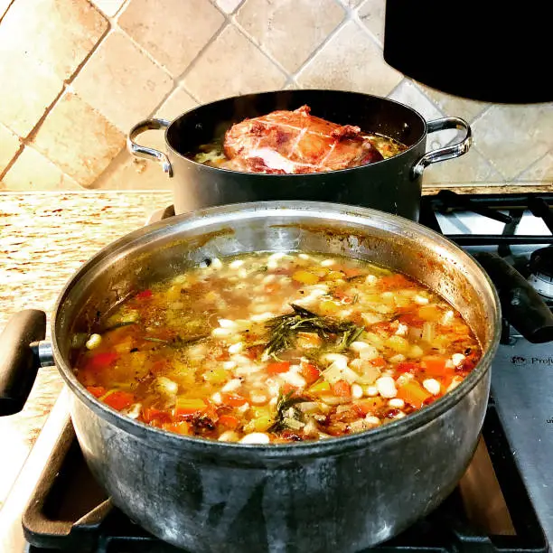 Two pots on the stove, 1 contains a ham hock and the second featuring a simmering soup of vegetables and bean
