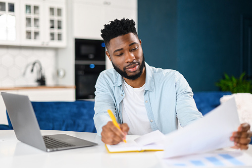Smart smiling African-American male student using laptop for studying online, taking education class, man preparing report working remotely from home, taking notes and looking through workpapers