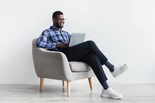 cuerpo entero de joven negro sonriente con computadora portátil sentado en sillón, trabajando en línea contra la pared blanca del estudio - red chairs fotografías e imágenes de stock