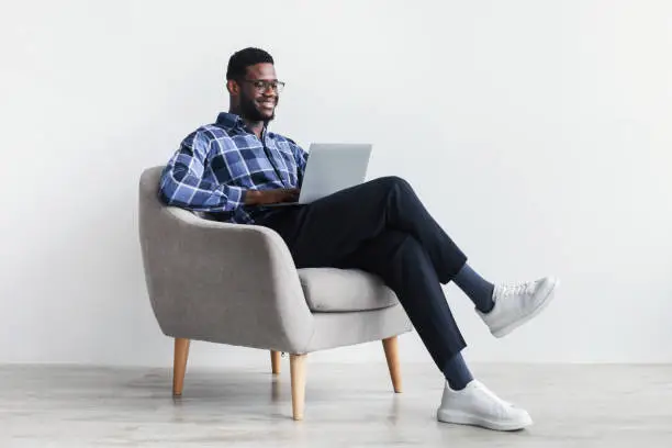 Photo of Full length of smiling young black guy with laptop computer sitting in armchair, working online against white studio wall