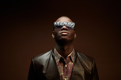 Portrait of African young man in stylish leather jacket and sunglasses posing on brown background