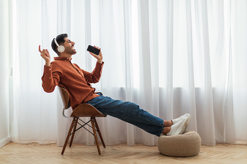 Carefree Singer. Cheerful young guy singing favorite song using cell phone as microphone, dancing wearing wireless headset. Excited man having fun at home sitting on chair, profile side view portrait