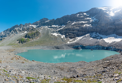 View of the turquoise-colored Griesslisee lake, the glacier cliff directly on the lake and the surrounding mountains. black boulders cover the glacier surface. Located near the famous mountain pass road Klausenpass in the canton of Uri.