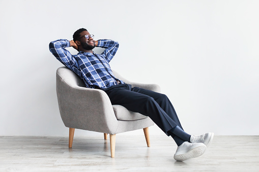 Peaceful young black man relaxing in armchair against white studio wall, free space
