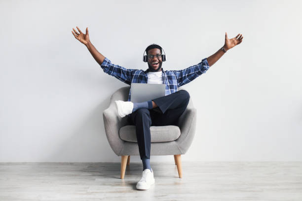 Excited African American guy in headphones greeting someone while chatting online on laptop, sitting in armchair Excited millennial African American guy in headphones gesticulating, greeting someone while chatting online on laptop, sitting in armchair against white wall, full length. Distance communication excitement laptop stock pictures, royalty-free photos & images