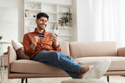 Chico árabe sonriente usando teléfono y tarjeta de crédito en casa photo